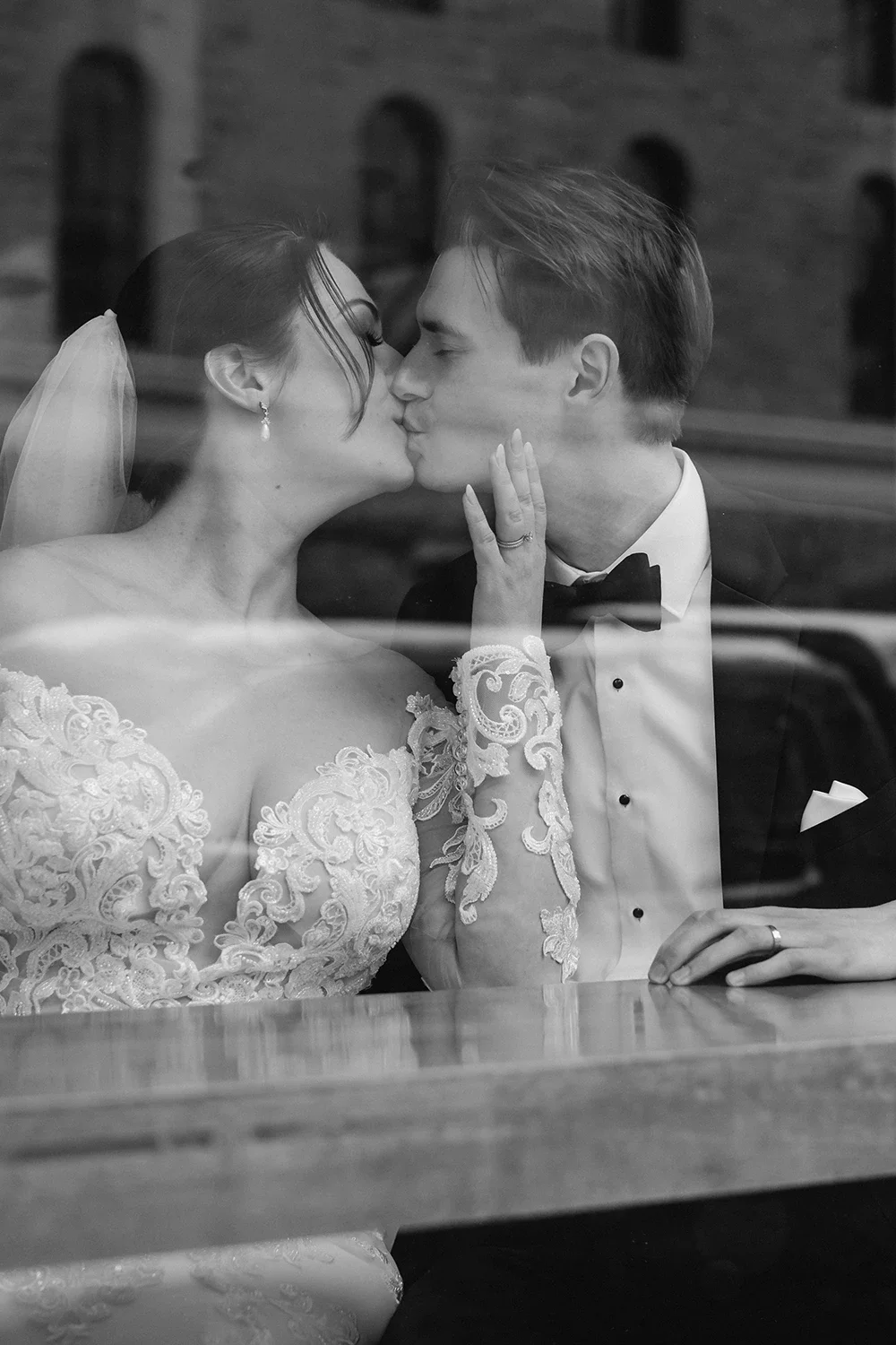 Black and white photo of a bride and groom sharing a kiss, viewed through a window. The bride wears an lace wedding dress and a veil, while the groom is in a tuxedo with a bow tie. The bride's hand with a wedding ring is on the groom's face.