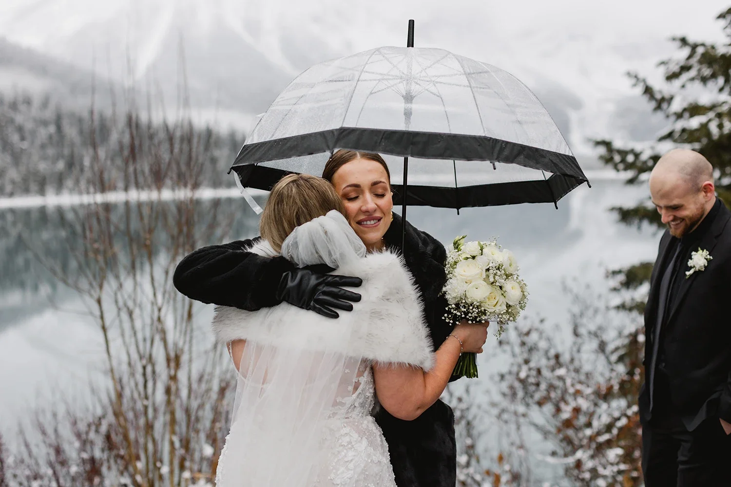 A woman in a wedding dress and a man in a suit sharing a hug under an umbrella outdoors during winter, with trees and snow in the background.
