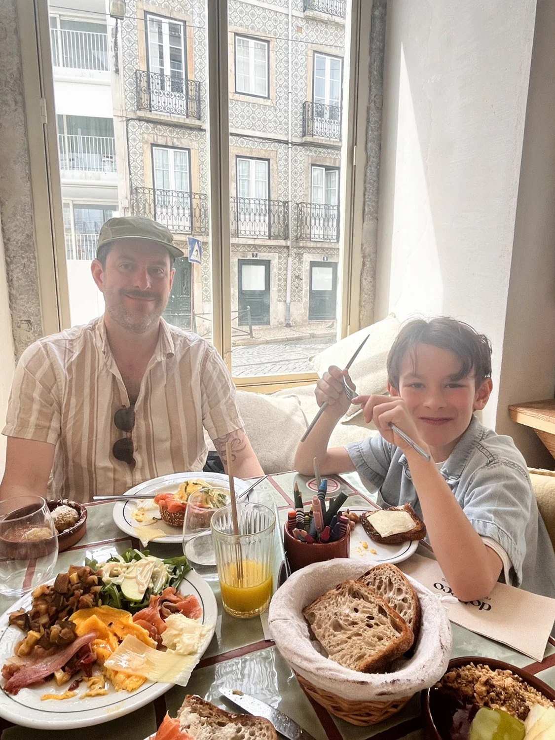 A man and a boy enjoying breakfast at a table with various food items, including bread, salad, and drinks, inside a restaurant with large windows showing a building outside.