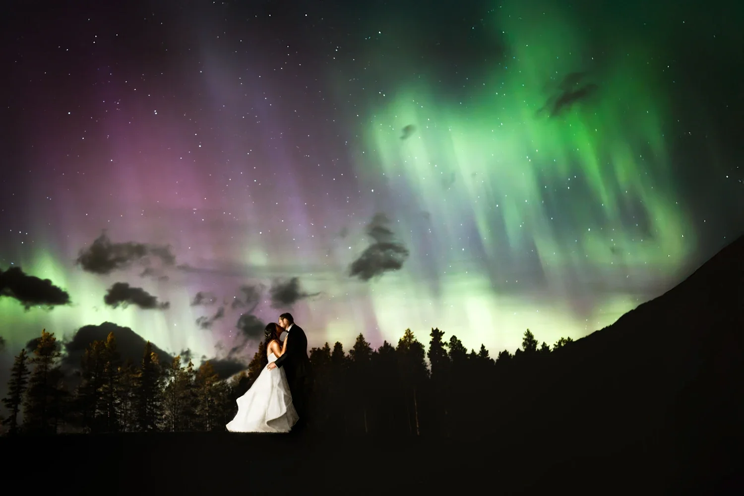 A couple dressed in wedding attire sharing a kiss outdoors at night, with the Northern Lights illuminating the sky in green and purple hues above them, silhouetted trees, and a mountain in the background.