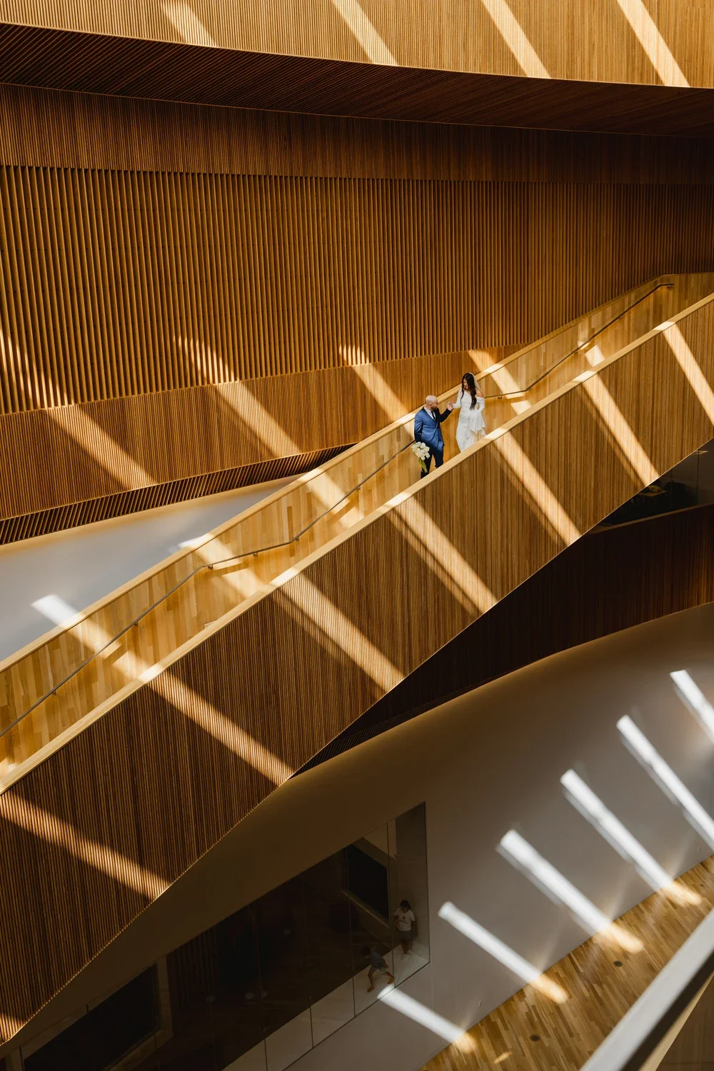 A couple dressed in wedding attire on a wooden staircase inside a modern building with geometric light patterns on the wall.