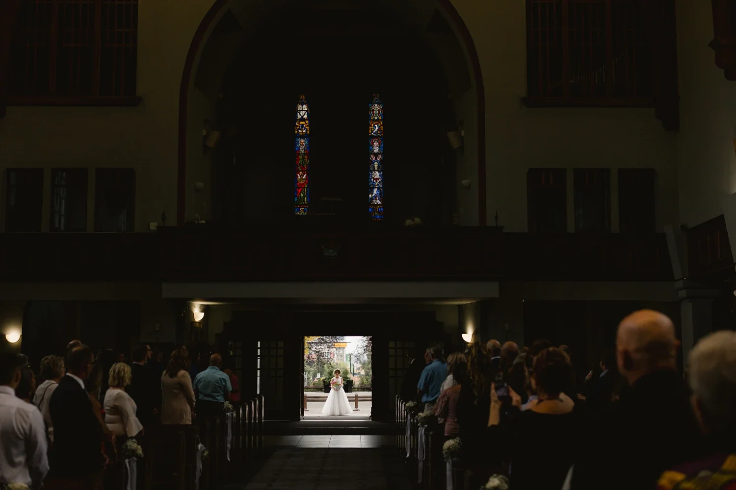 Wedding ceremony inside a church, with the bride standing at the altar in a white gown, guests seated on either side, and stained glass windows in the background.