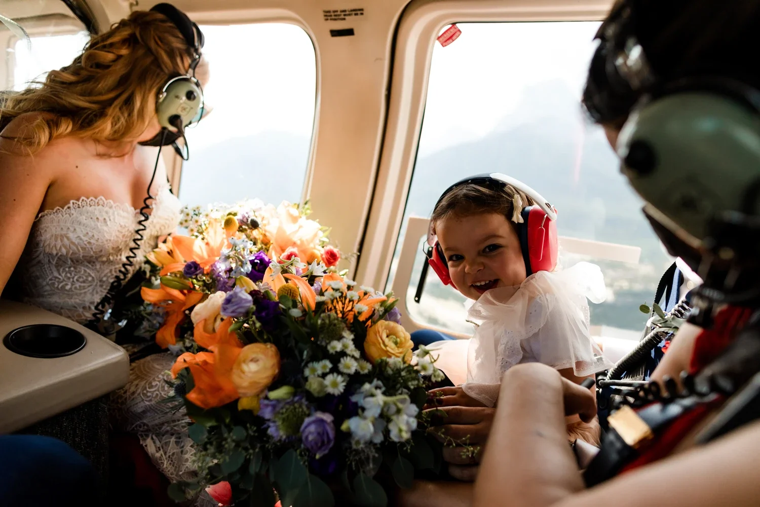 A woman, a young girl, and a man in an aircraft cabin with a large bouquet of flowers, the woman and girl wearing headsets, the girl smiling with a scrunched face, and the woman bending toward her.