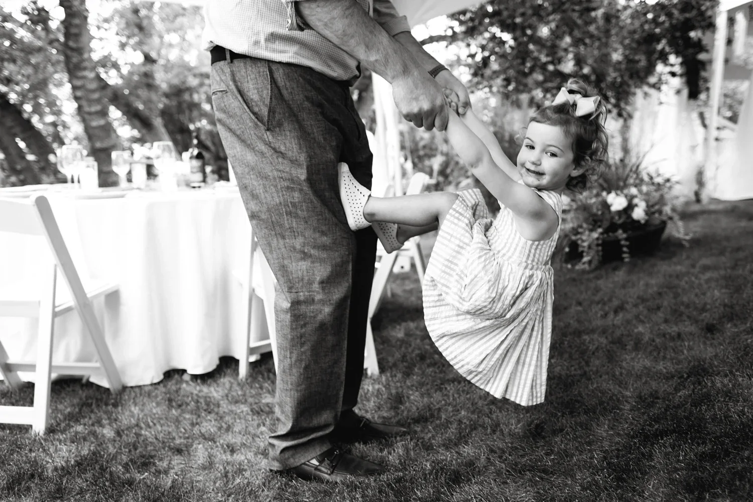 A young girl in a dress is lifting her foot while holding hands with an older man, outdoors in a garden setting, with a table with drinks and chairs in the background.