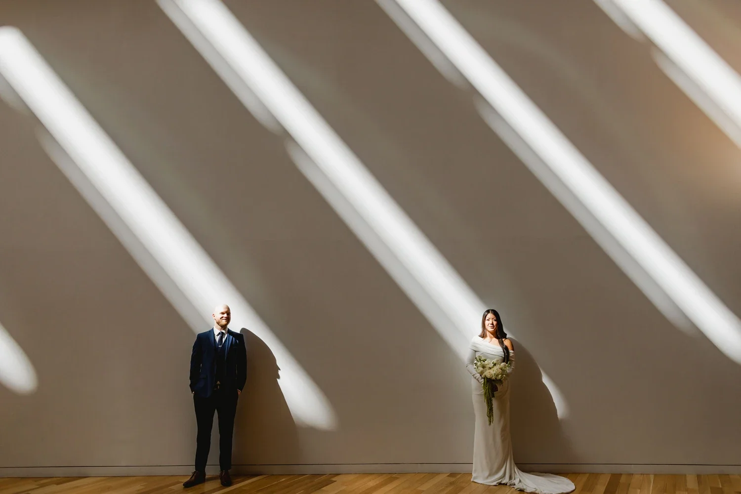 A man in a navy suit and a woman in a white wedding dress holding a bouquet, standing apart against a beige wall with large shadows cast by overhead lighting.
