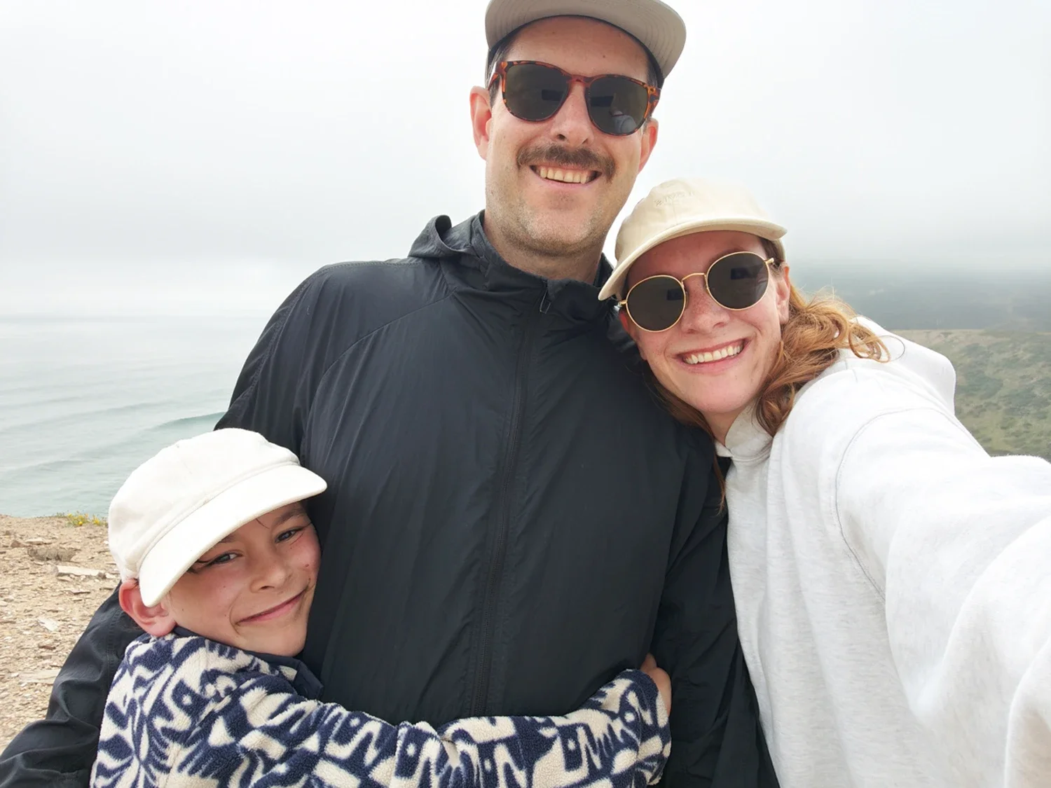 A family of three taking a selfie outdoors near a coastal area with cliffs and ocean in the background. The mother, father, and child all wear hats and sunglasses, smiling warmly.