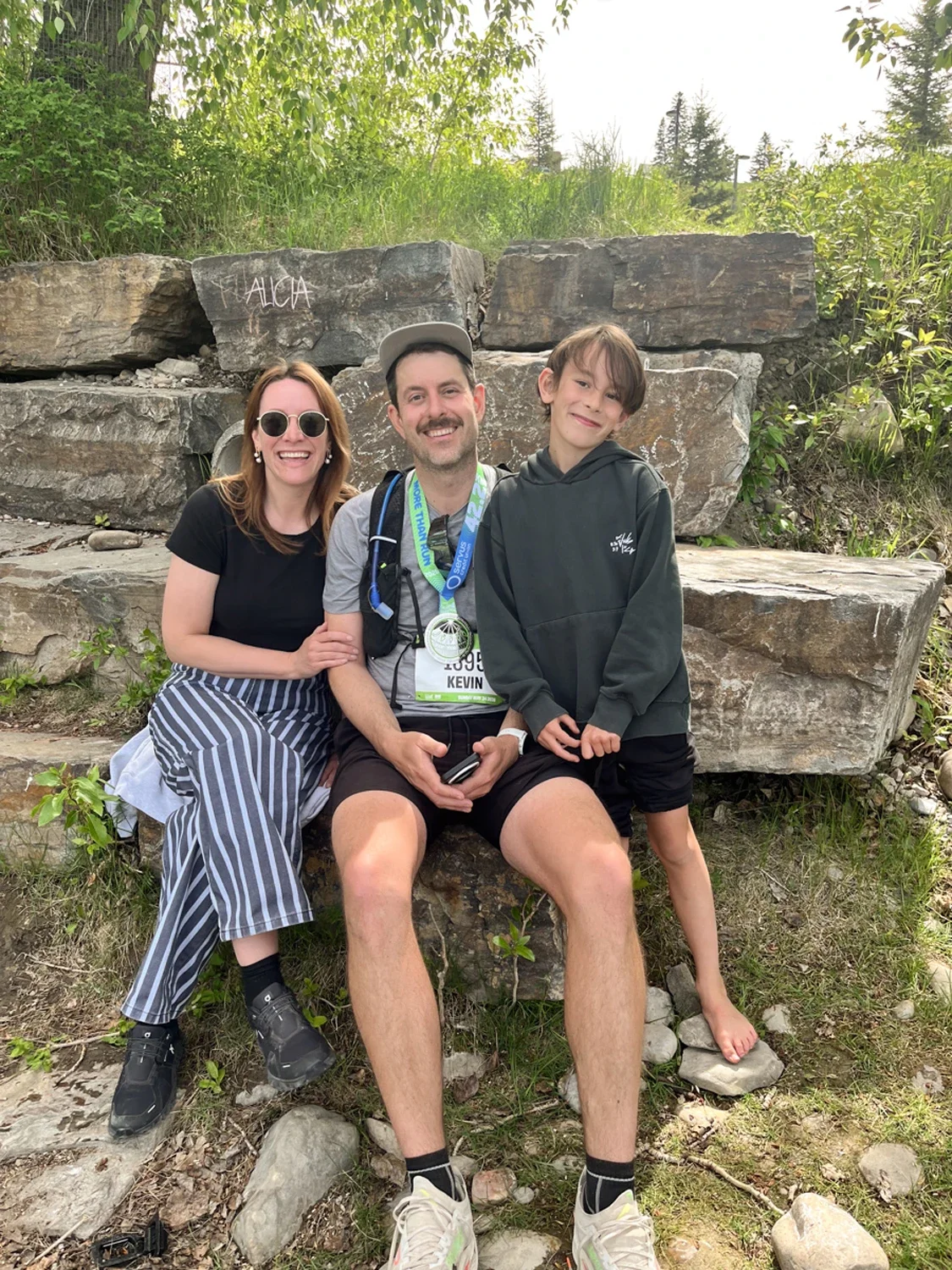 A woman, a man, and a boy sitting on rocks outdoors with green trees behind them. The woman is wearing sunglasses and a black shirt with striped pants. The man has a medal around his neck, a gray hat, and holds a phone. The boy is wearing a gray hood
