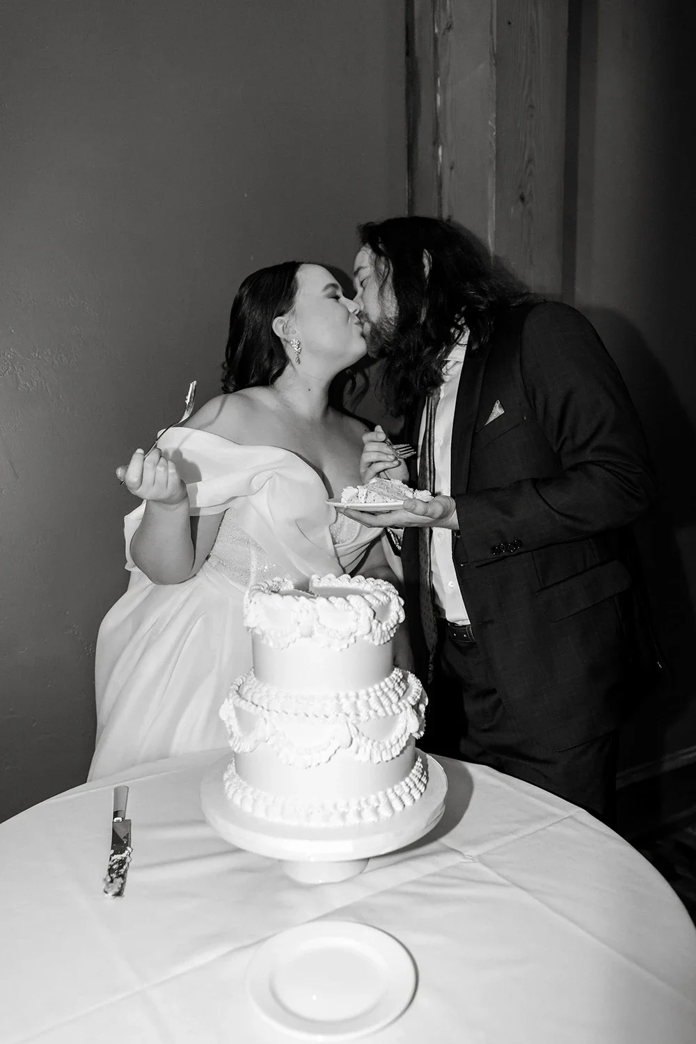 A couple sharing a kiss at their wedding reception, with a wedding cake and slices of cake on a table in front of them.