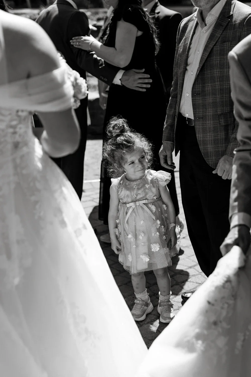A young girl with curly hair wearing a dress with floral patterns, standing among adults during a social event, possibly a wedding, on a sunny day.