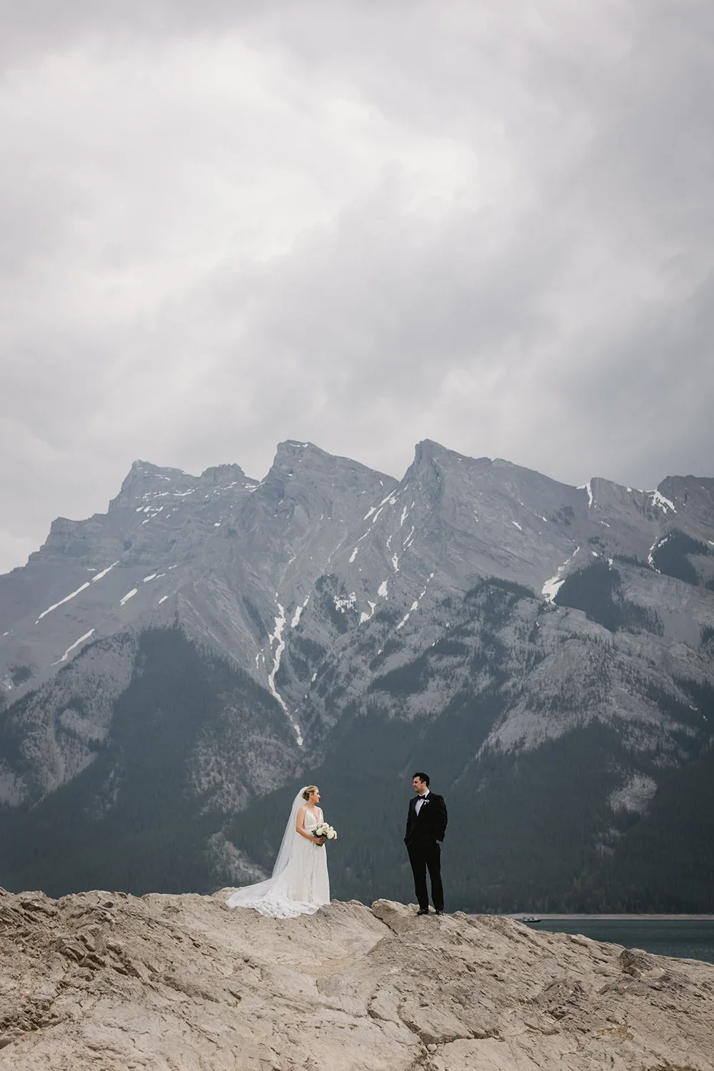 A bride and groom stand on rocky terrain in front of a mountainous landscape with cloudy sky.