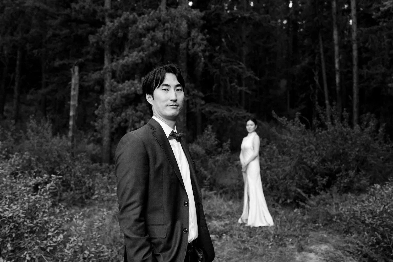 A man in a tuxedo standing outdoors in the foreground, with a woman in a white gown standing in the background among trees and shrubs, in a black-and-white photograph.