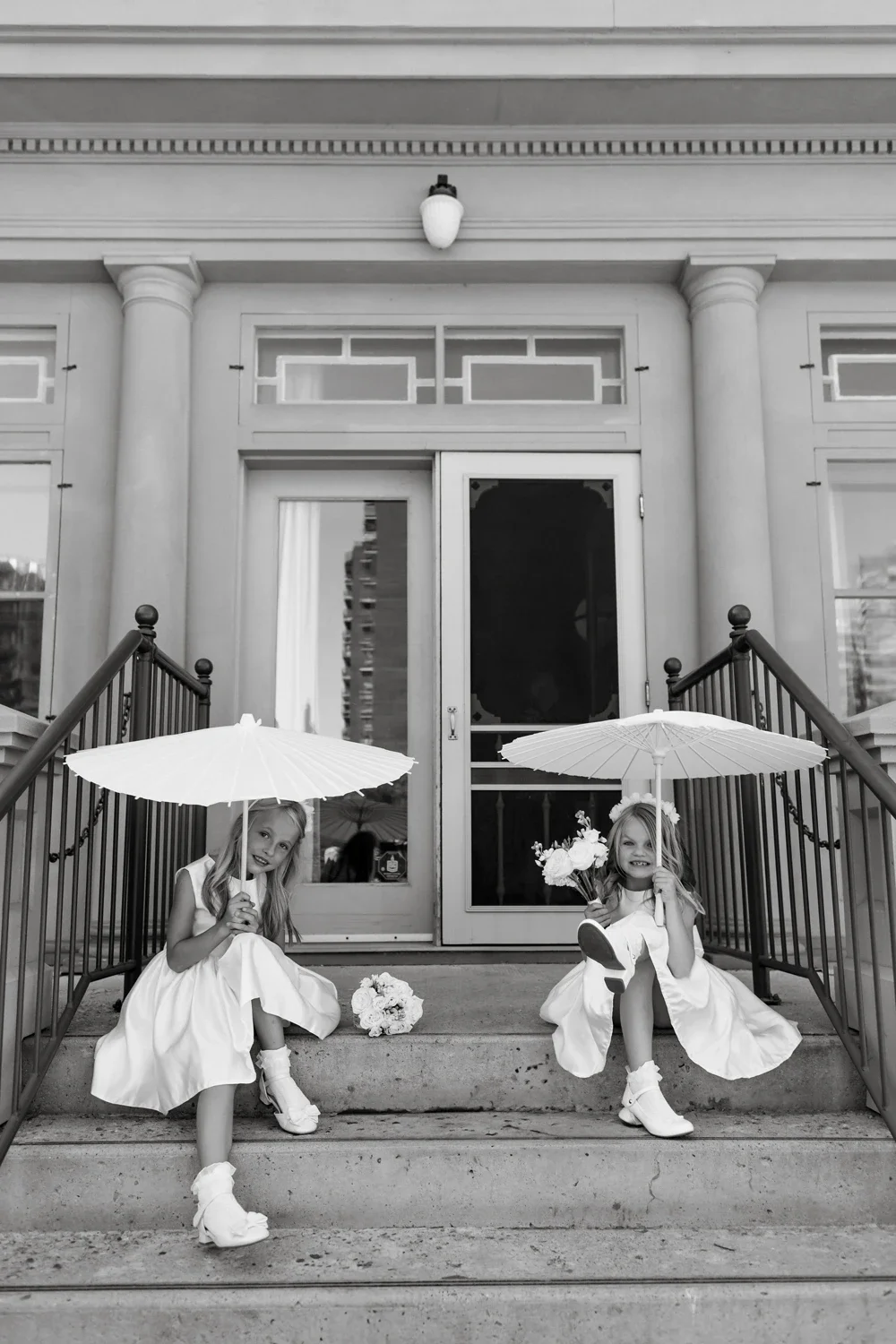 Two young girls in white dresses sit on steps outside a building with columns, holding umbrellas and flowers, smiling.