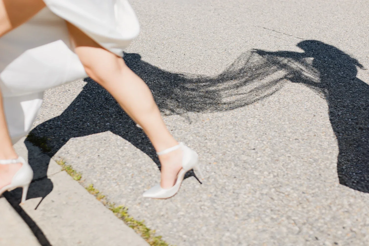 A woman in white high heels and a white dress crouching down on the sidewalk, casting a shadow that resembles a person in a suit and fedora hat.