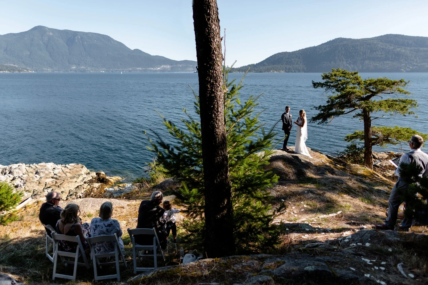 A wedding ceremony taking place outdoors by a lake with mountains in the background. The bride and groom are standing on a rocky area, exchanging vows, with a few guests seated nearby, and officiant on the right side.