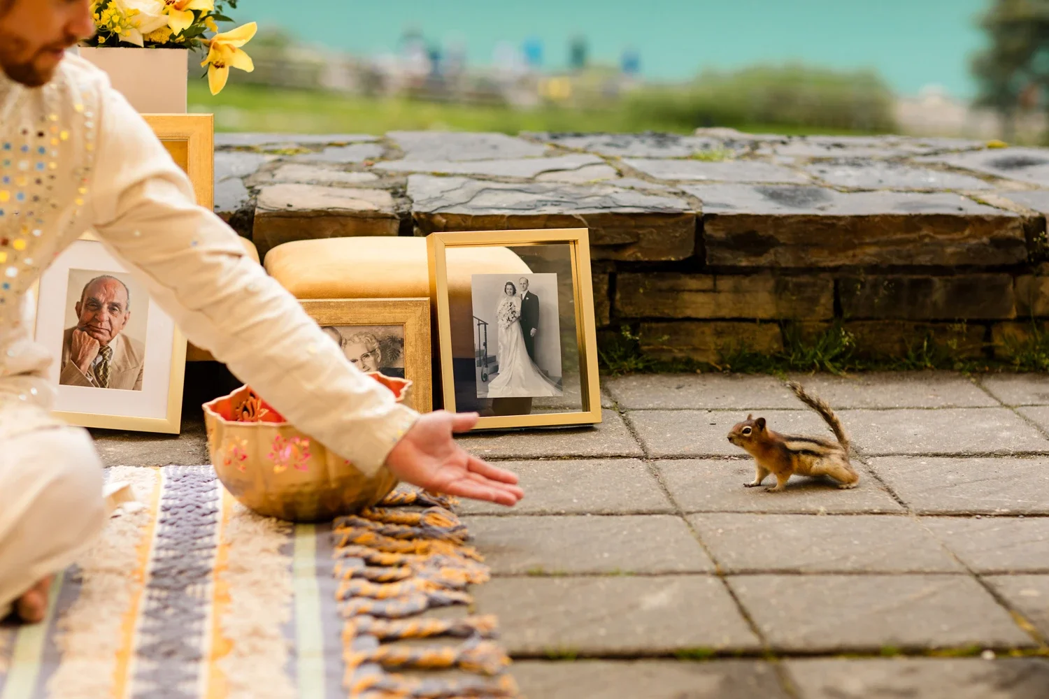 Person participating in a prayer or meditation session outdoors, sitting on a colorful mat with framed photographs of elderly and wedding couples behind them, and a small squirrel on the paved ground nearby.