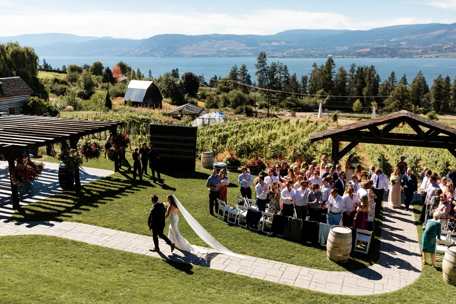 Outdoor wedding ceremony with a bride and groom walking along a paved path, surrounded by guests, vineyards, and scenic mountains under a blue sky.