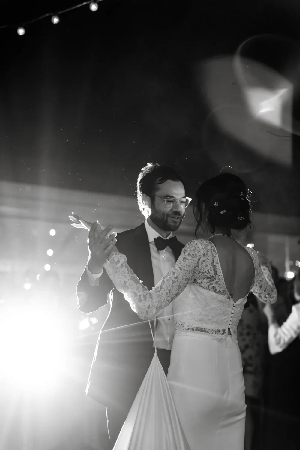 A couple, dressed in wedding attire, sharing a dance at their wedding reception in a dimly lit venue with stage lighting and other guests in the background.