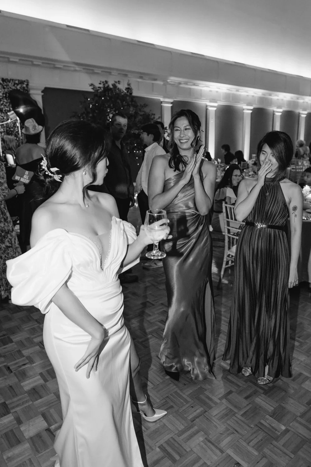 Three women in elegant gowns are laughing and chatting at a formal event, one holding a glass of wine.