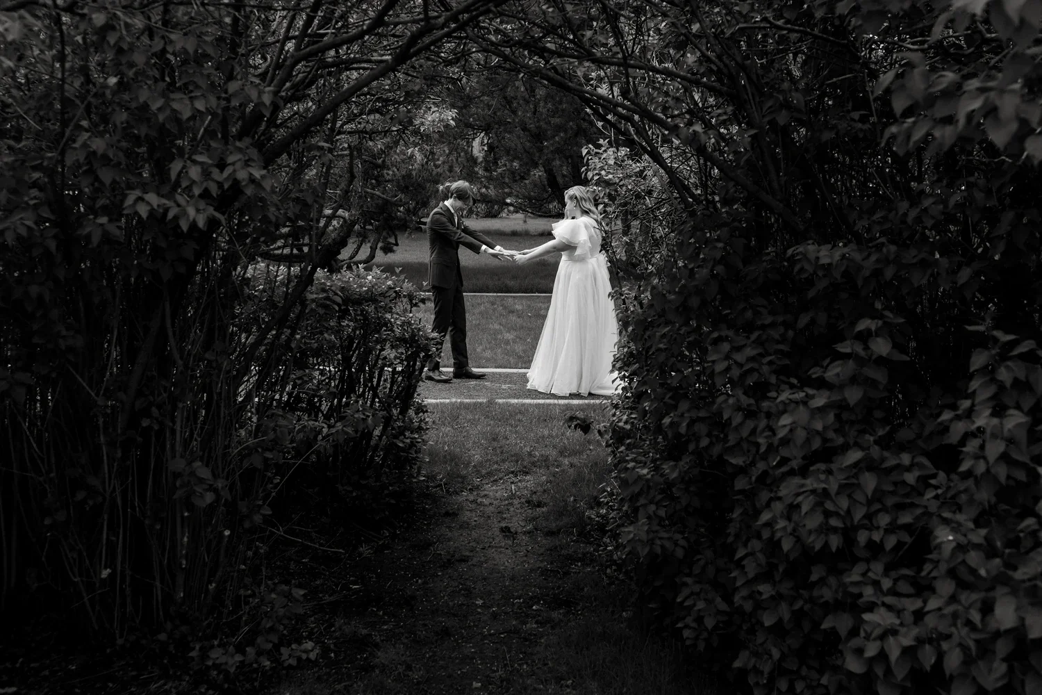 A black and white photo of a bride and groom during their wedding ceremony, holding hands and exchanging vows outdoors between garden bushes.