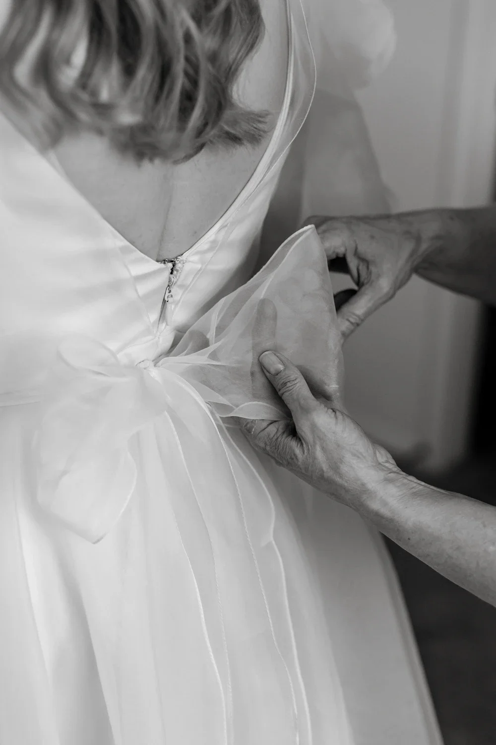 An elderly woman helps a younger person put on a wedding dress.