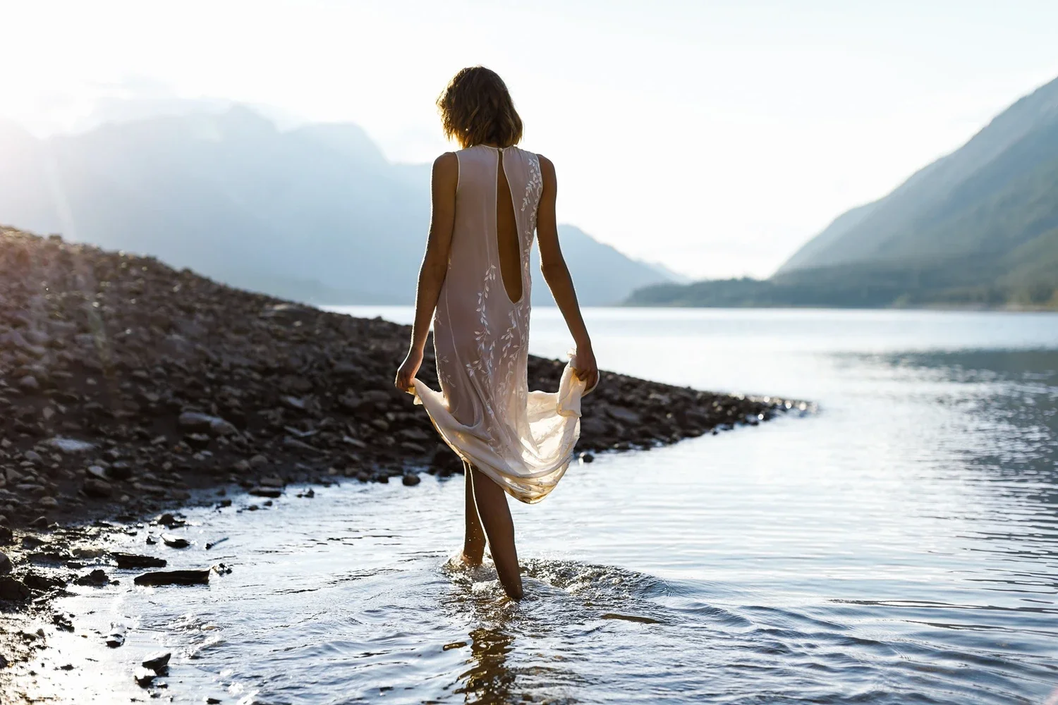 A woman in a light-colored dress walking in a river near a rocky shoreline, with mountains in the background and the sun setting or rising.