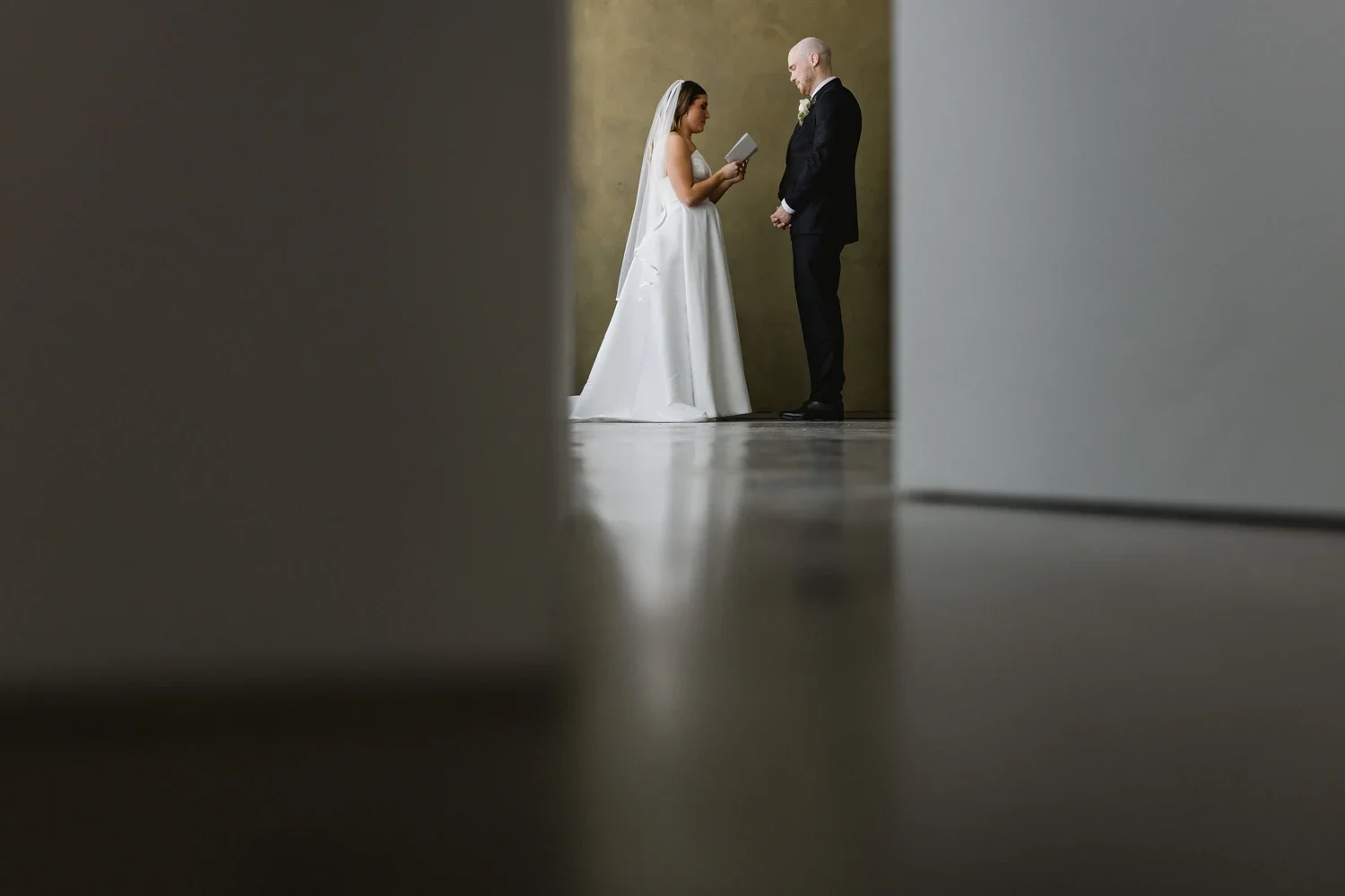 A bride and groom standing and exchanging vows during their wedding ceremony, viewed through a narrow opening in a wall.