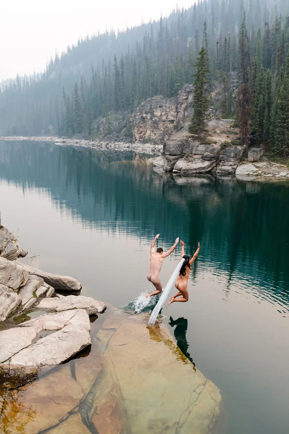 A man and woman jumping off a rock into a mountain lake surrounded by pine trees and rocky cliffs.