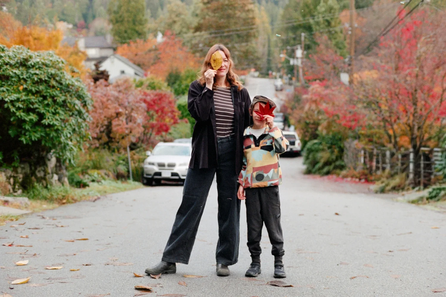 A woman and a boy standing on a street in autumn, both holding colorful leaves in front of their faces. The woman is wearing a black jacket and wide-leg pants, the boy is wearing a patterned hoodie and dark pants. The street is lined with trees with 