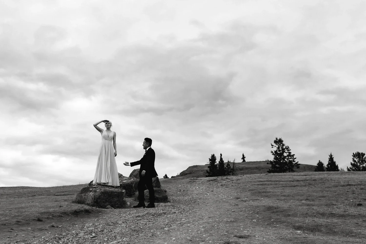 A black and white photo of a man in a suit standing on the ground and a woman in a long dress standing on a large rock, with a cloudy sky and trees in the background.