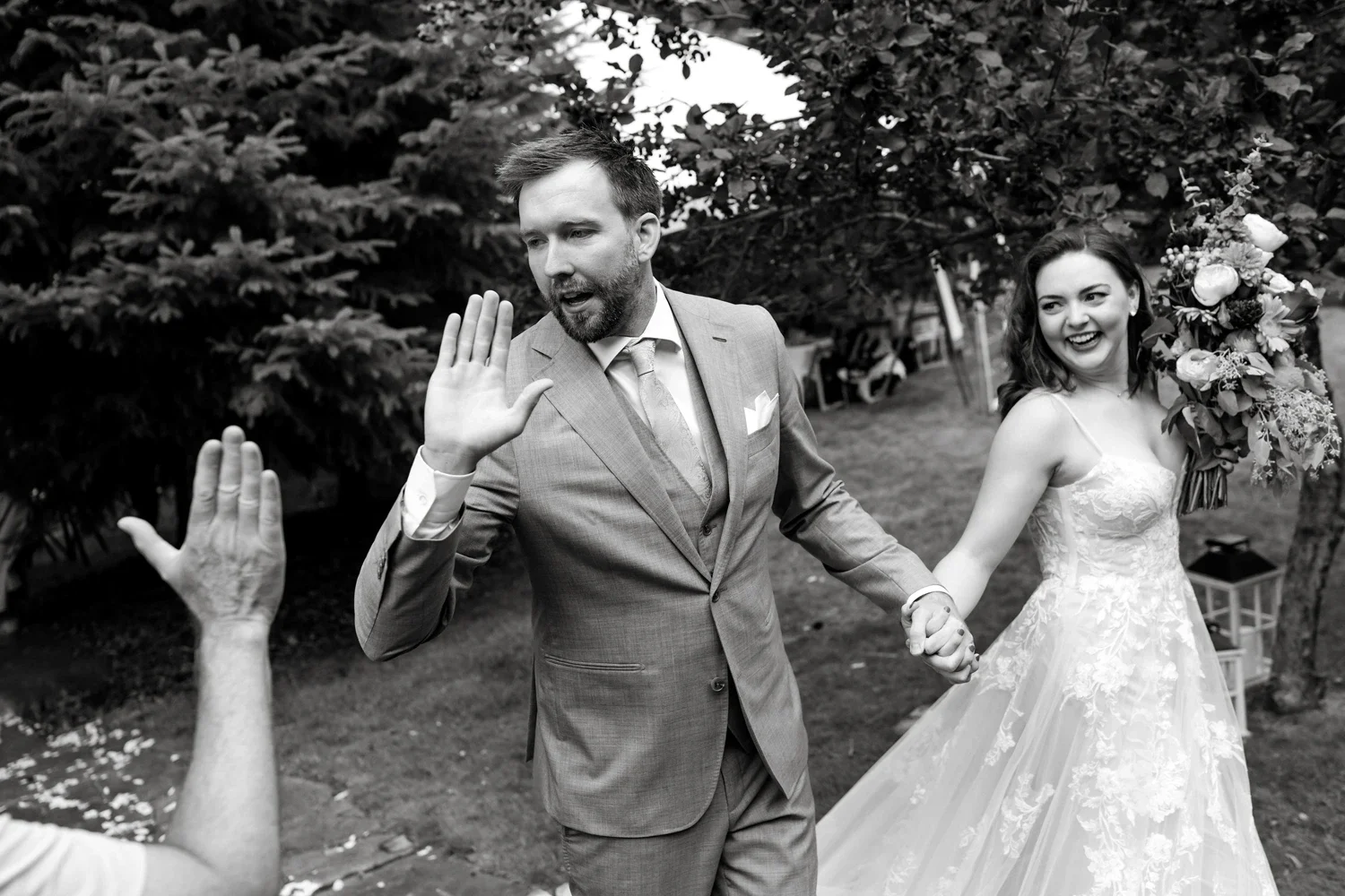 A wedding scene with a bride and groom holding hands, walking outdoors. The groom is giving a high-five to someone off-camera, and the bride is smiling, holding a large bouquet of flowers.