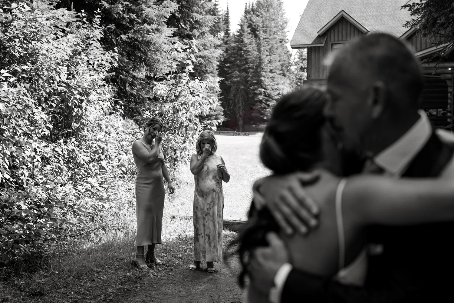 A couple is embracing and hugging each other, with the woman smiling and the woman's eyes closed, in focus. Two women in dresses are standing in the background near a wooded area, embracing and wiping tears from their faces, likely emotional or cryin