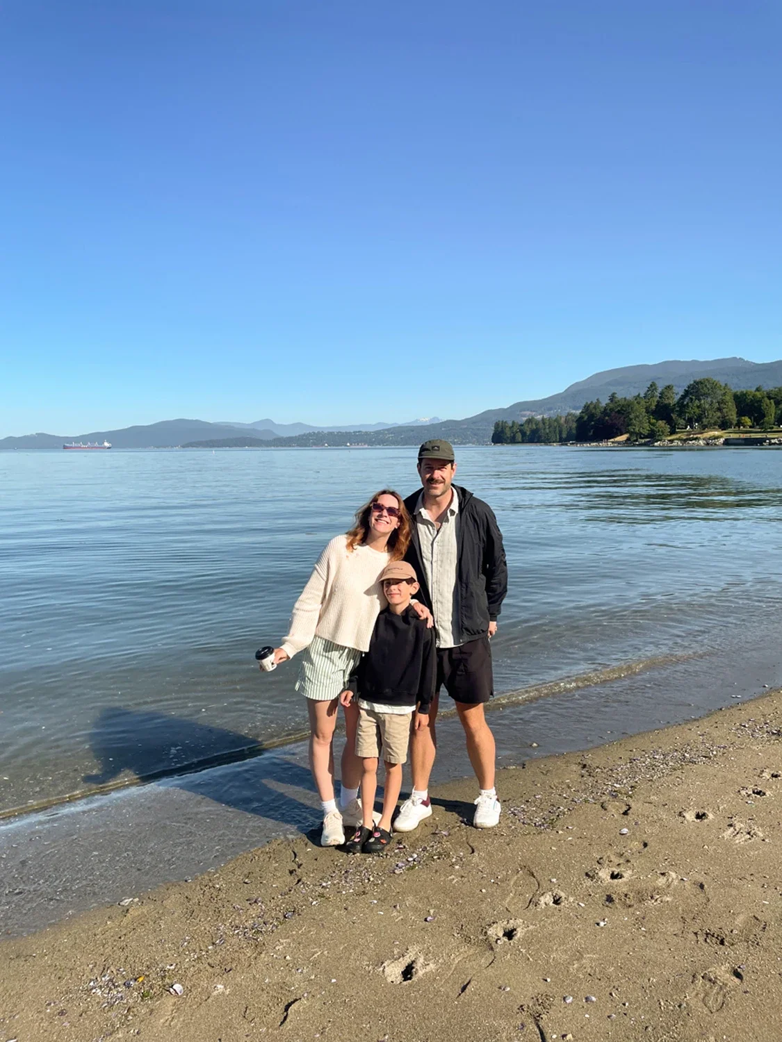 A family of three standing on a sandy beach by calm water, with mountains and trees in the background on a clear, sunny day.