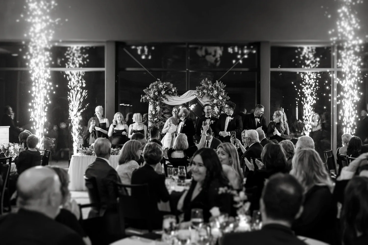 Black and white photo of a wedding celebration with the bride and groom kissing under a floral arch. Guests seated at tables watch and cheer, with some taking photos. Sparkler effects are visible on either side of the scene.