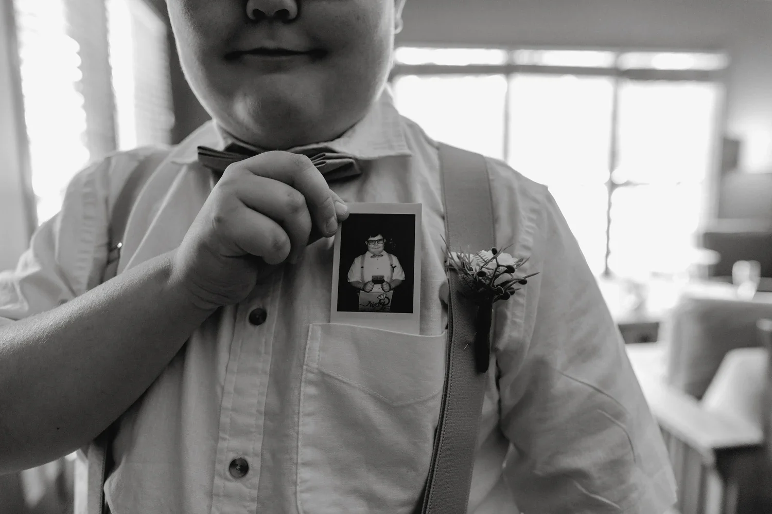 A person dressed in a white shirt with a bow tie, holding a small photograph of themselves in front of their chest. The person has suspenders and a boutonnière pinned to their shirt. The photo appears to be taken in a bright room with large windows.