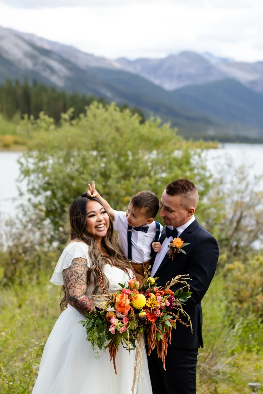 A joyful bride, groom, and young child celebrating outdoors near a lake with mountains in the background. The bride holds a colorful bouquet, and the groom has a boutonniere. The child is making a peace sign.