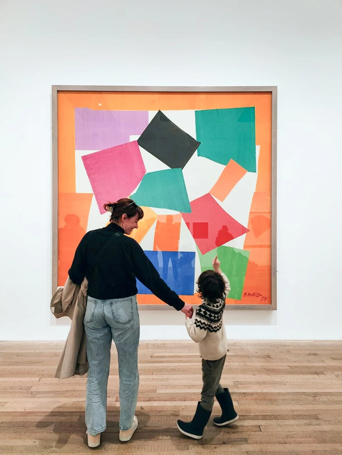 A woman and a young girl are in an art gallery looking at a large colorful abstract painting of geometric shapes on the wall. The woman is holding the girl's hand while gesturing towards the artwork.