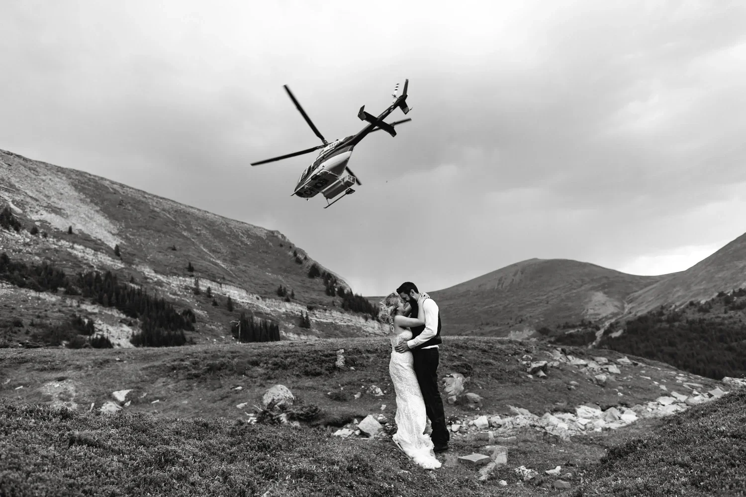 A couple in wedding attire embracing in a mountainous landscape with a helicopter flying overhead.