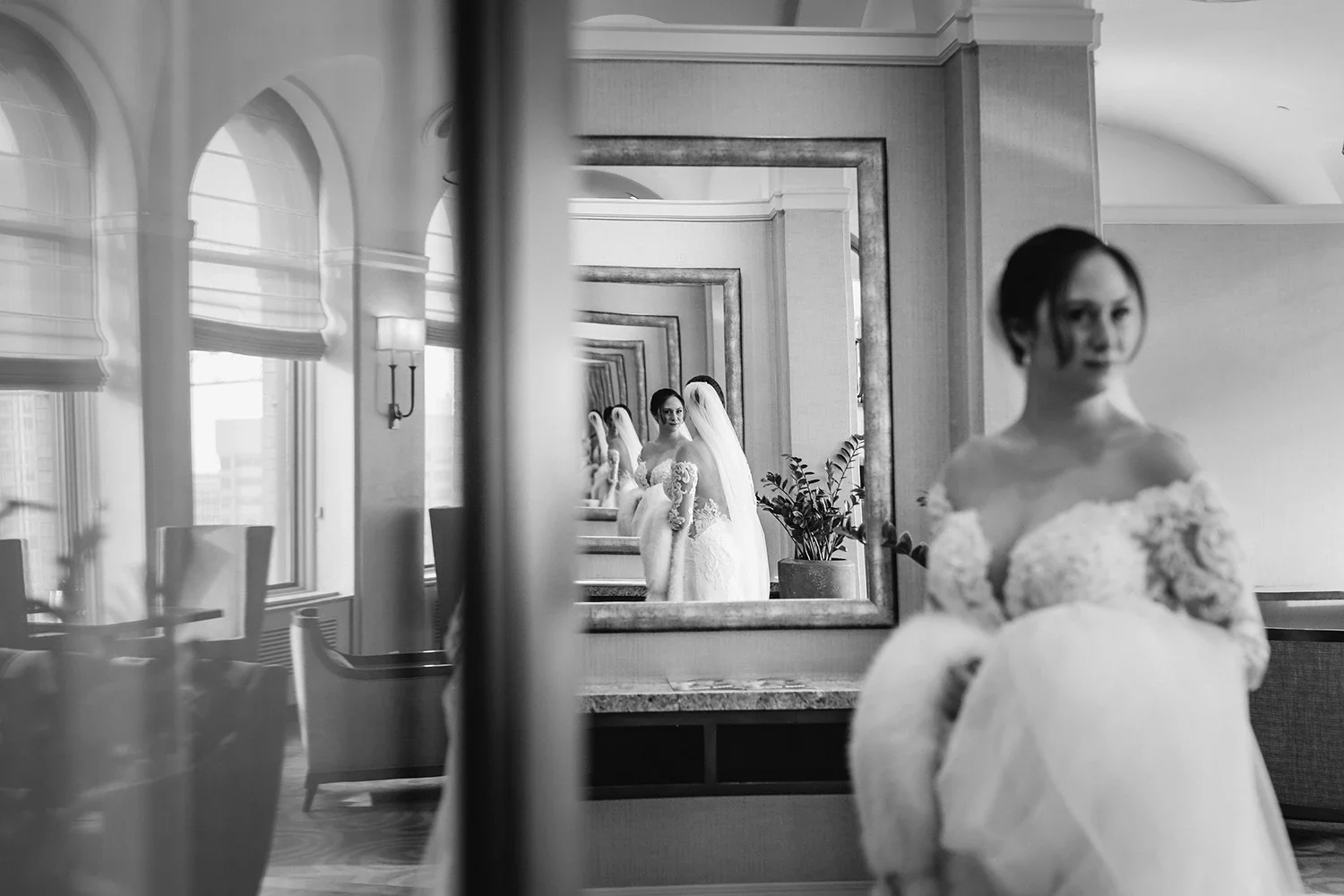 A black-and-white photo of a bride looking at herself in a large mirror. The bride is wearing a wedding dress with lace details and a veil. The mirror reflects her image multiple times, creating an illusion of depth. The room has large windows with s