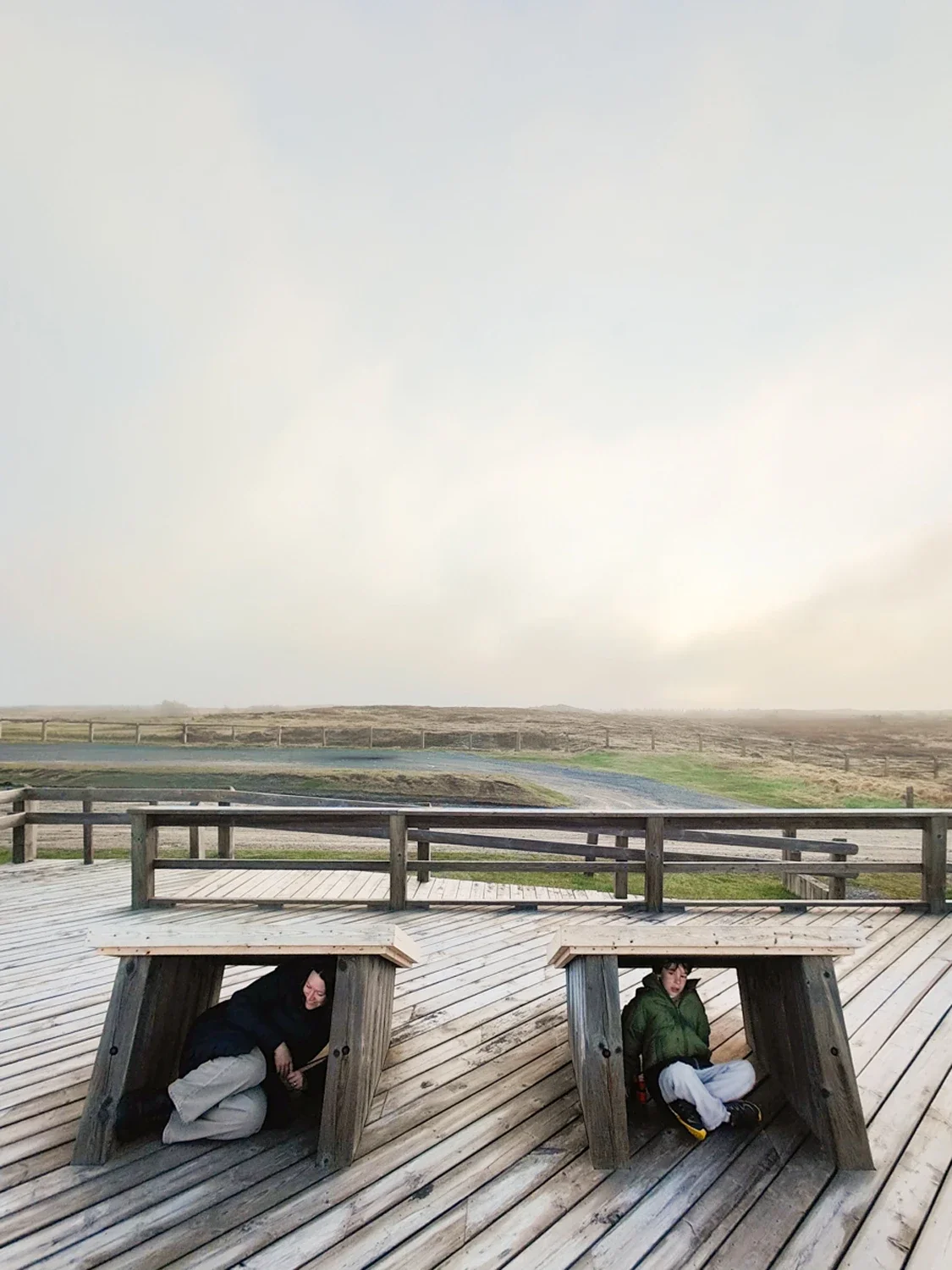 Two people sitting under small wooden structures on a wooden deck outdoors, with open landscape and a cloudy sky in the background.