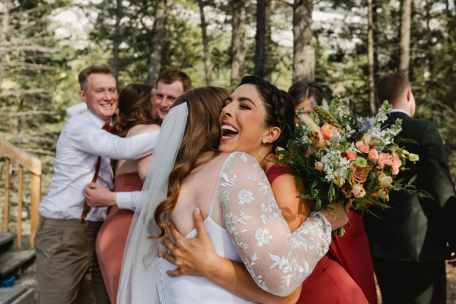 Bride hugging another woman with a bouquet, other guests in the background hugging and smiling outdoors.