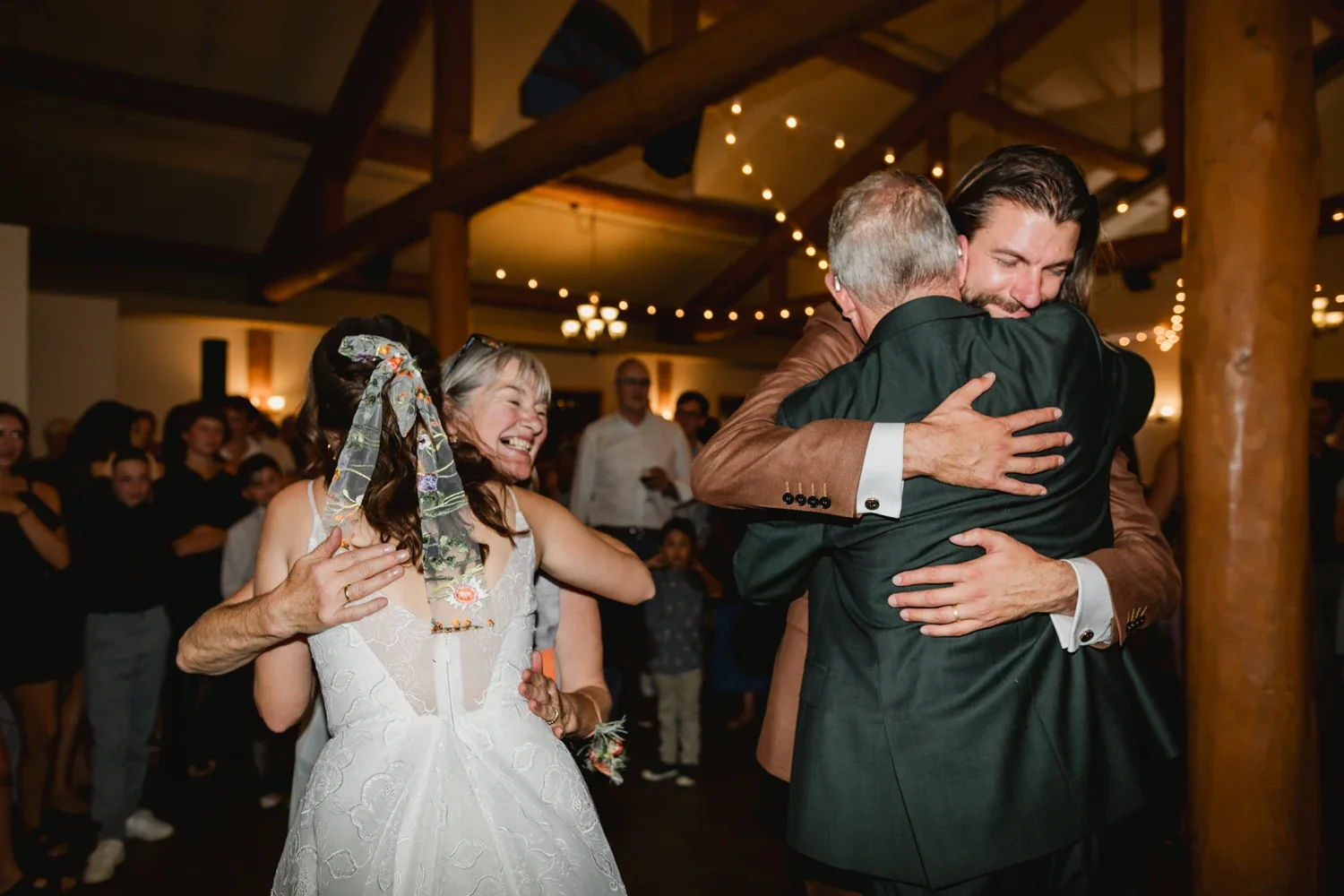 People celebrating at a wedding reception, with two men hugging and a woman laughing behind them.