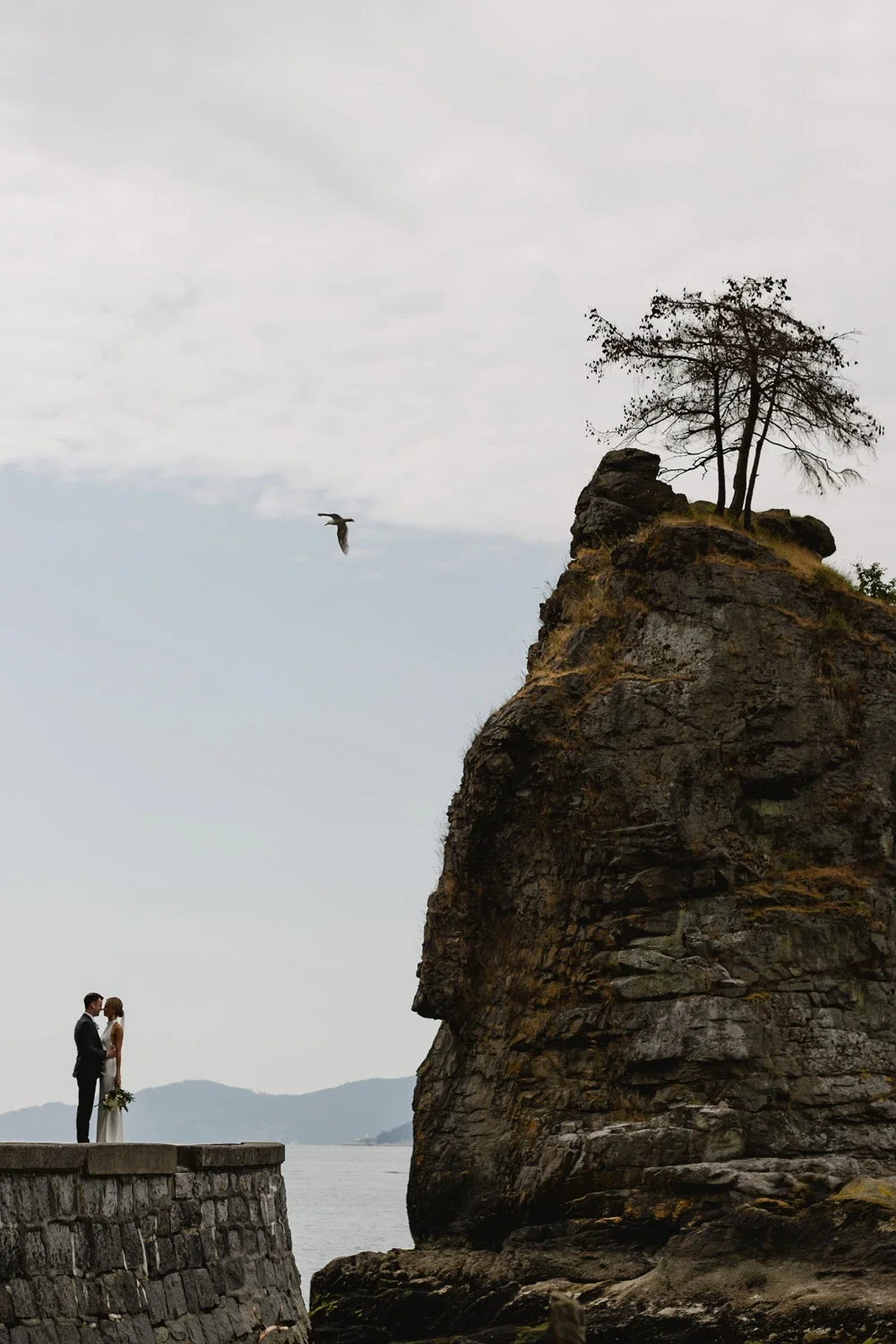 A couple in wedding attire standing on a stone ledge by water, facing each other, with a large rocky cliff with sparse trees and a bird flying overhead in the background.