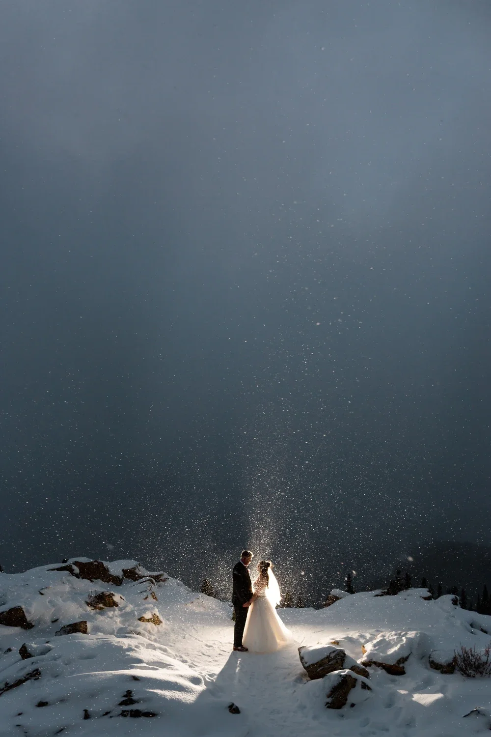 A couple in wedding attire stands on a snowy landscape at night, surrounded by falling snow, with a dark sky above.