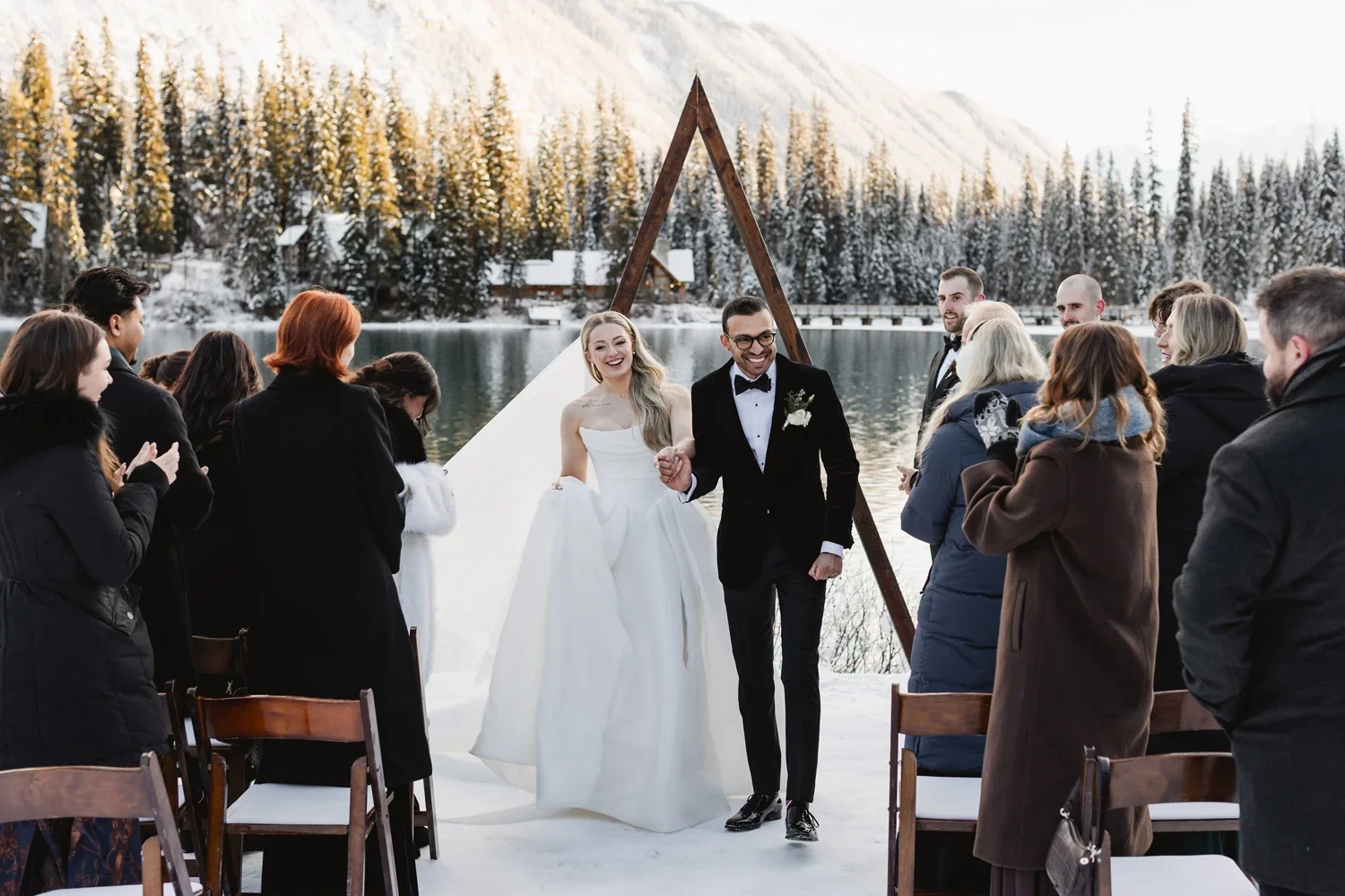 A wedding ceremony outdoors in a snowy landscape, with a bride and groom walking hand in hand, surrounded by guests, near a lake with snow-covered trees and mountains in the background.