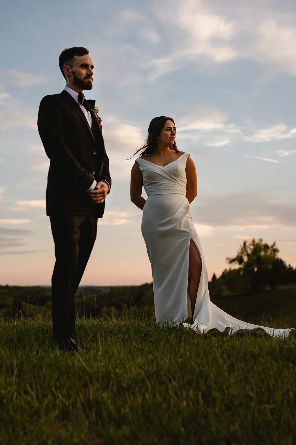 A bride and groom stand outdoors on grass during sunset, facing slightly away from each other, with a cloudy sky and distant trees in the background.