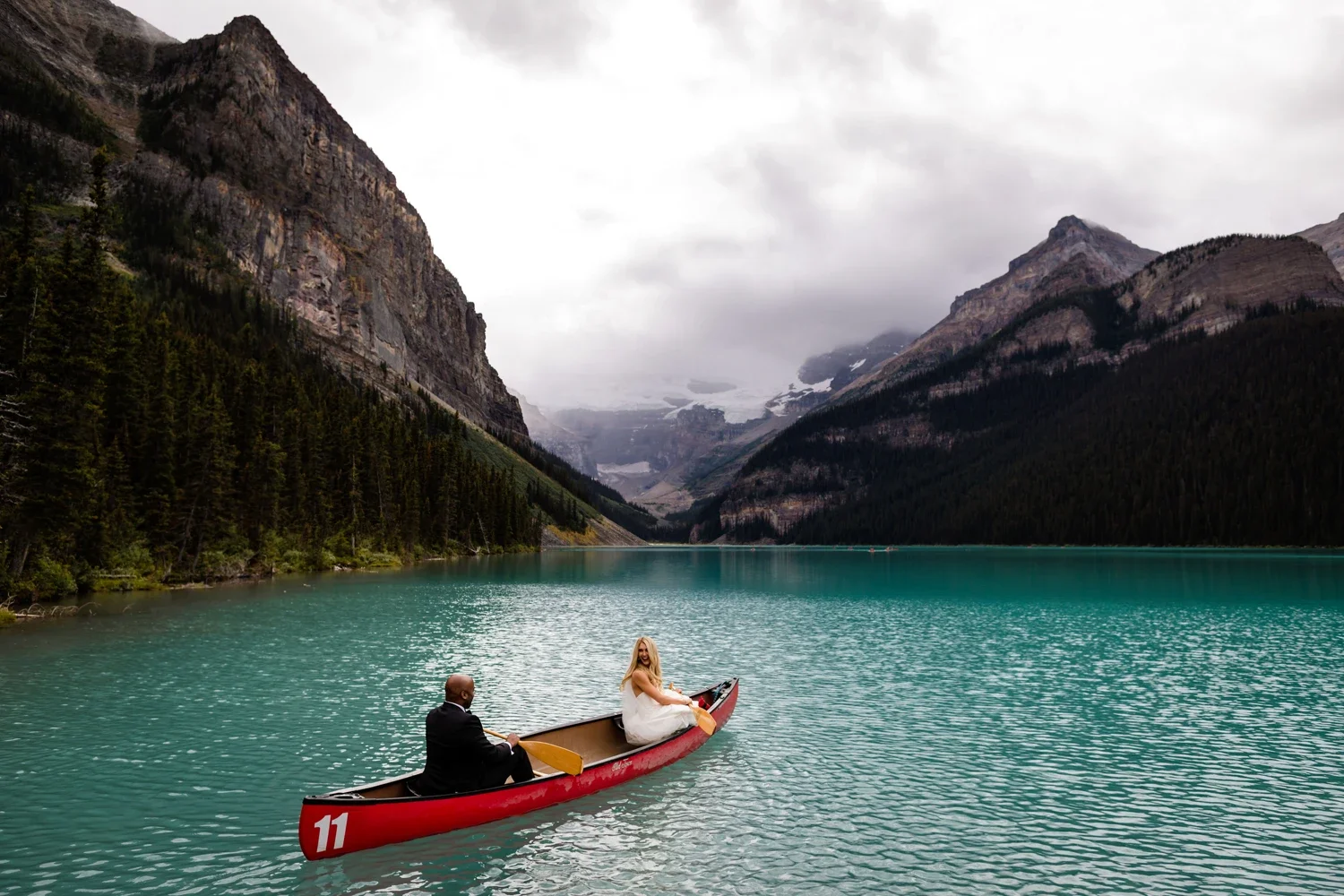 A couple in wedding attire row a red canoe on a turquoise lake surrounded by tall mountains and forested slopes under an overcast sky.