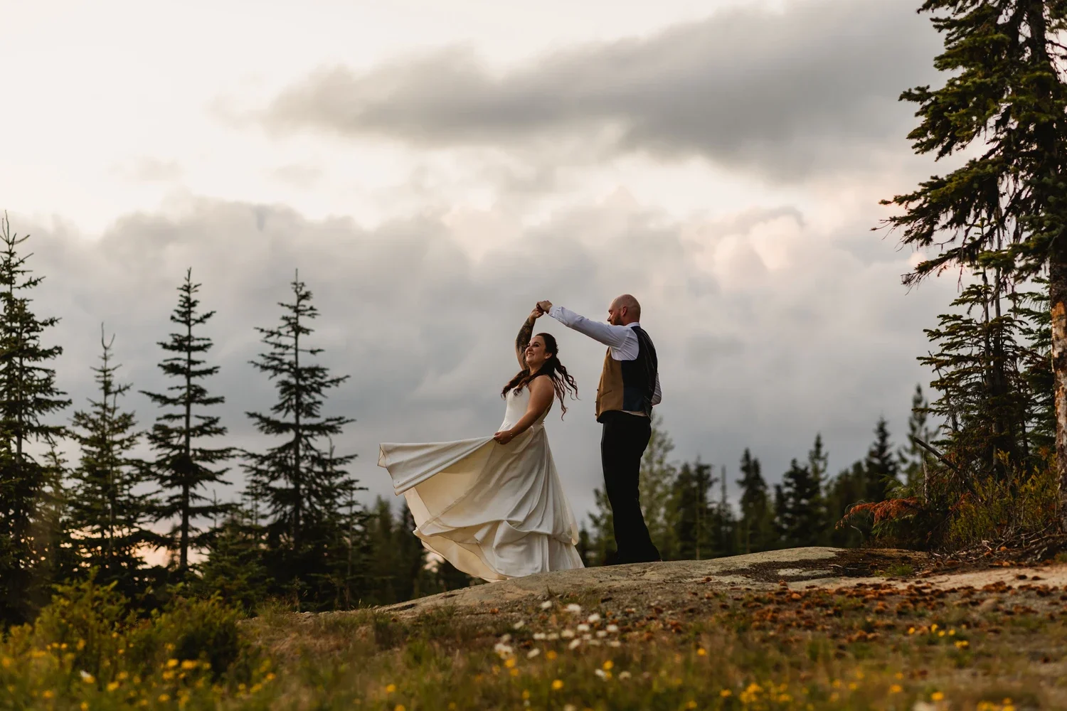 A couple dancing outdoors on a grassy hill with tall pine trees and a cloudy sky in the background, during sunset or early evening.