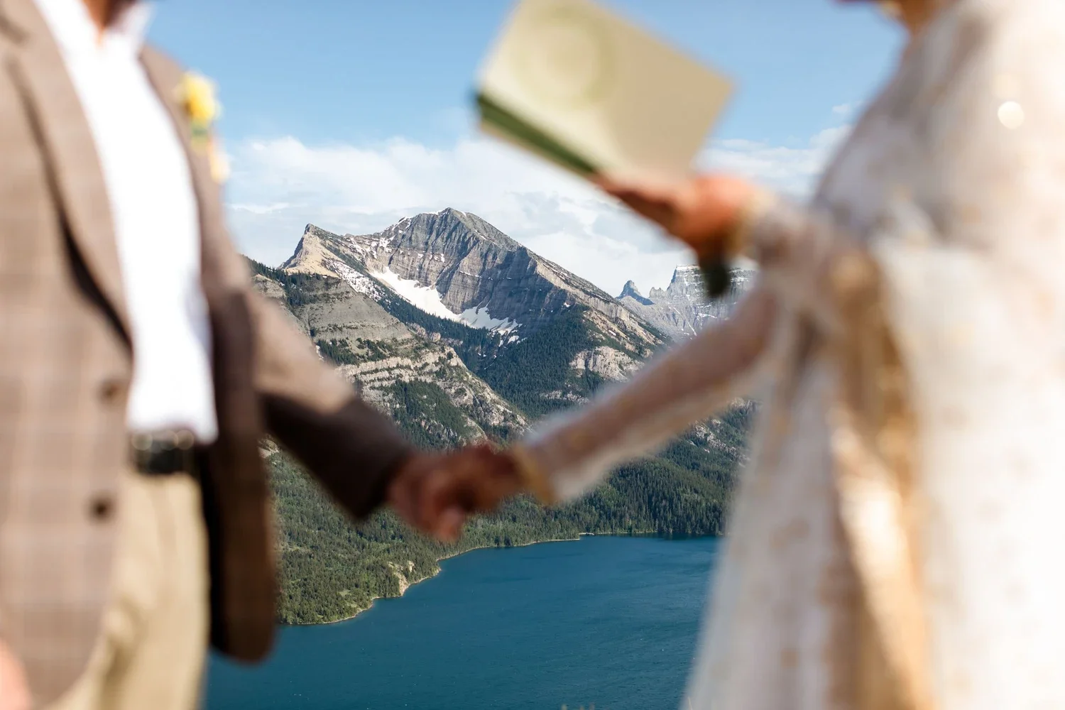 Blurred image of a couple holding hands with a mountain and lake in the background, the couple appears to be getting married or celebrating.