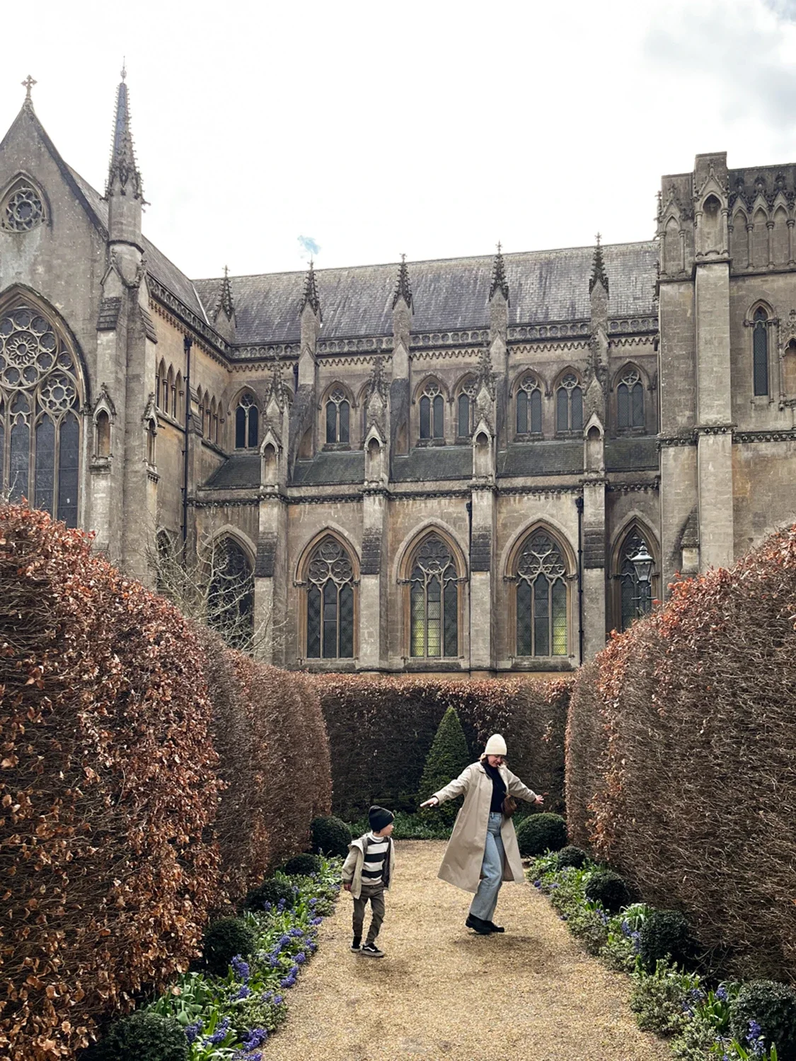 A woman and a child walking through a garden with blooming flowers and well-trimmed bushes in front of a large Gothic-style cathedral with stained glass windows.