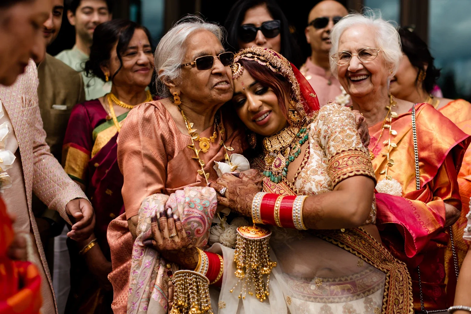 Indian bride with traditional jewelry hugging an elderly woman, surrounded by family members at a wedding celebration.